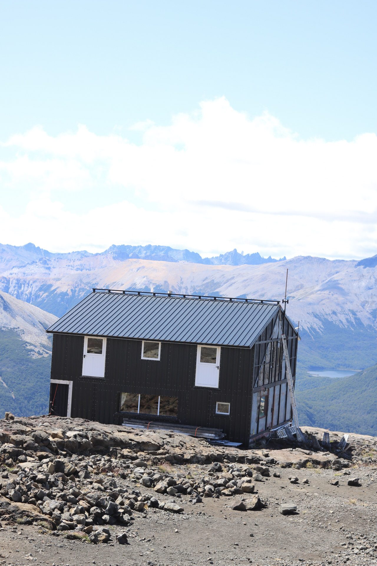 Refúgio rústico de madeira com telhado metálico escuro no cume do Cerro Catedral, em Bariloche, com montanhas nevadas e lago ao fundo