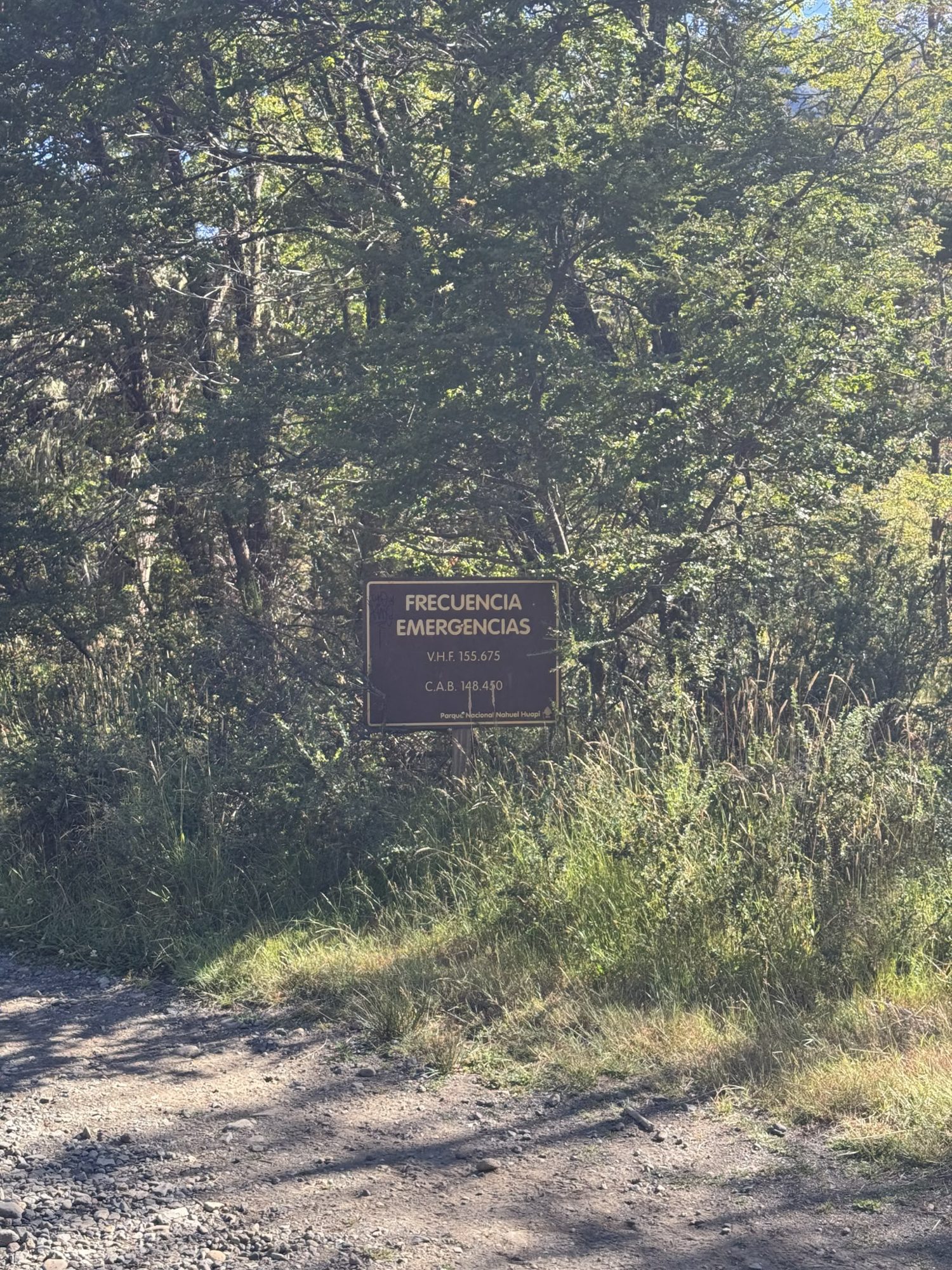 Placa marrom com frequências de emergência VHF 155.675 e CAB 148.450 do Parque Nacional Nahuel Huapi, próximo à trilha de o abrigo de montanha, cercada por vegetação nativa patagônica