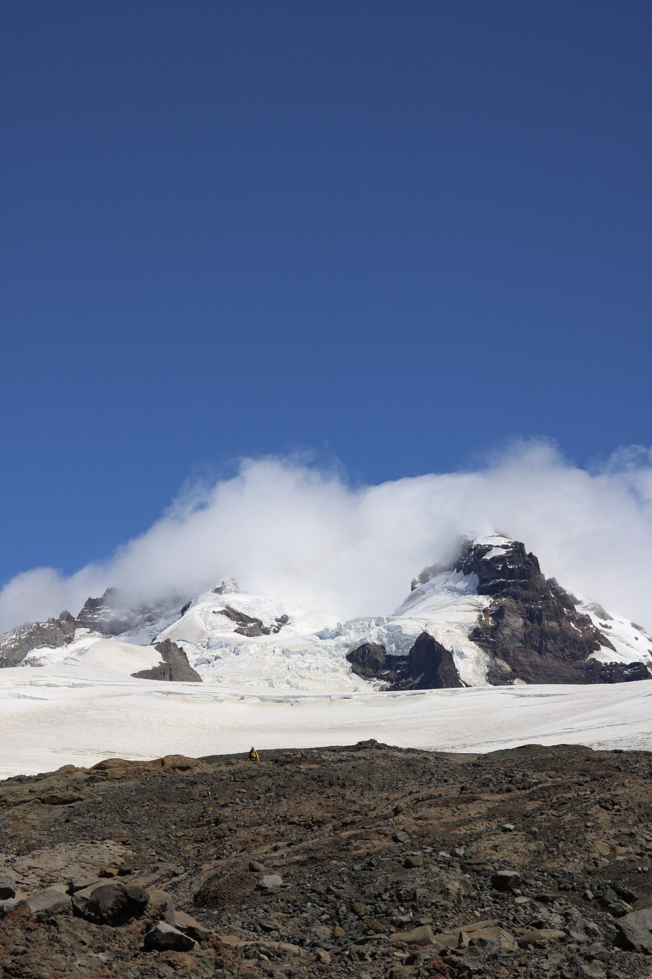 Glaciar patagônico em Bariloche com pico rochoso coberto de neve e gelo azul, nuvens envolvendo o cume e alpinista minúsculo ao fundo em agosto