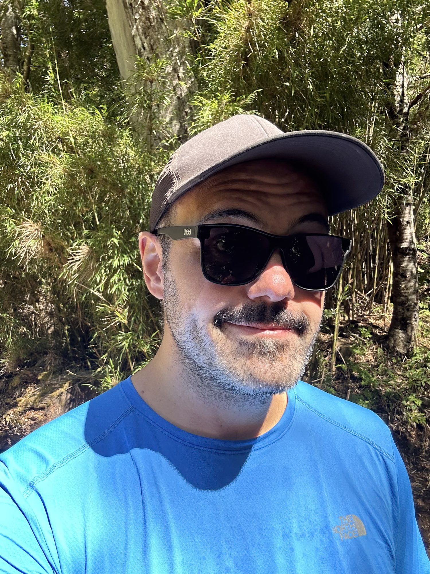 Homem com boné cinza, óculos de sol e camiseta azul da The North Face fazendo selfie em trilha cercada de vegetação em o abrigo de montanha, Patagônia argentina