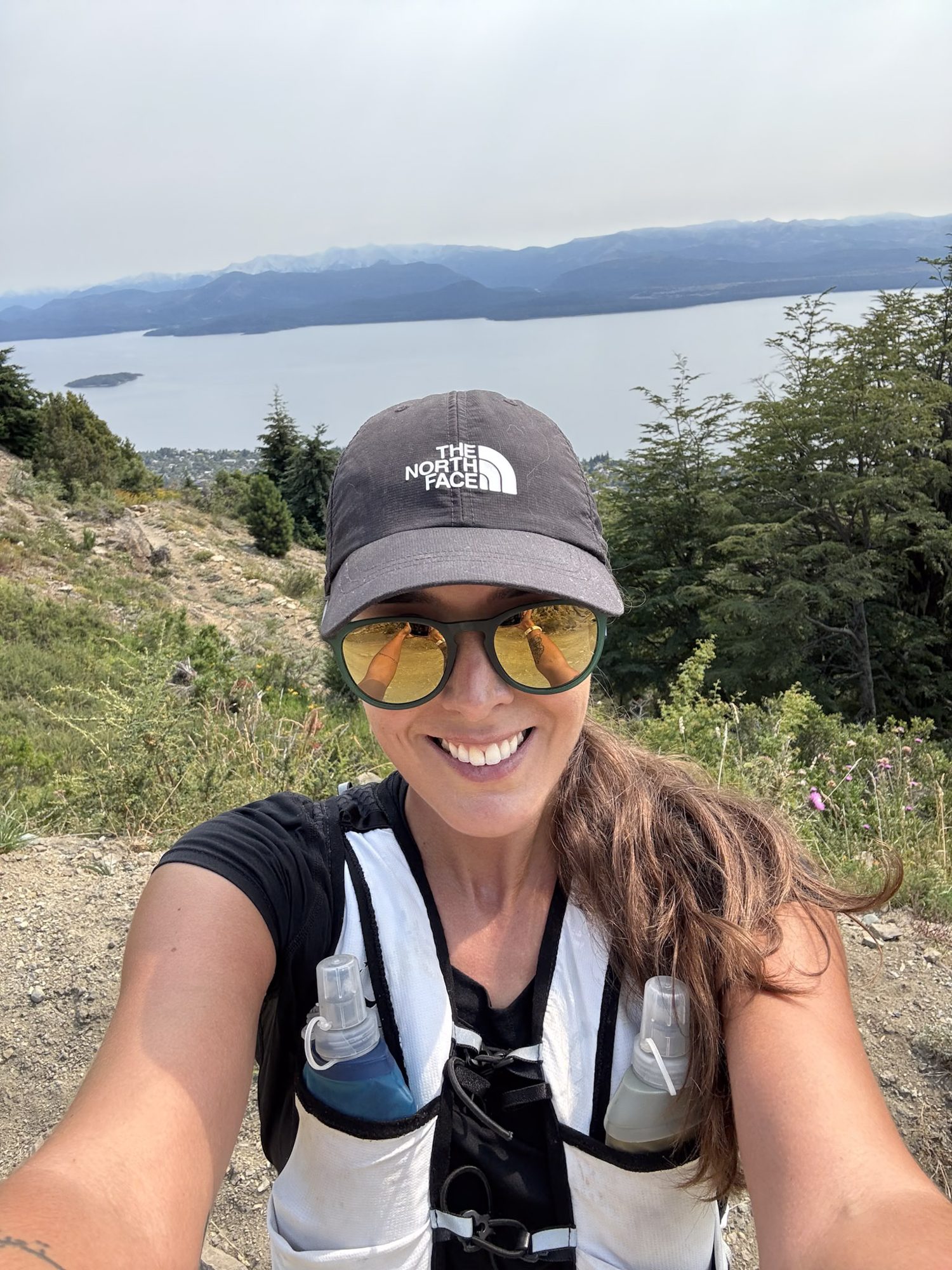 Corredora de trilha sorri em selfie com lago e montanhas ao fundo em Bariloche