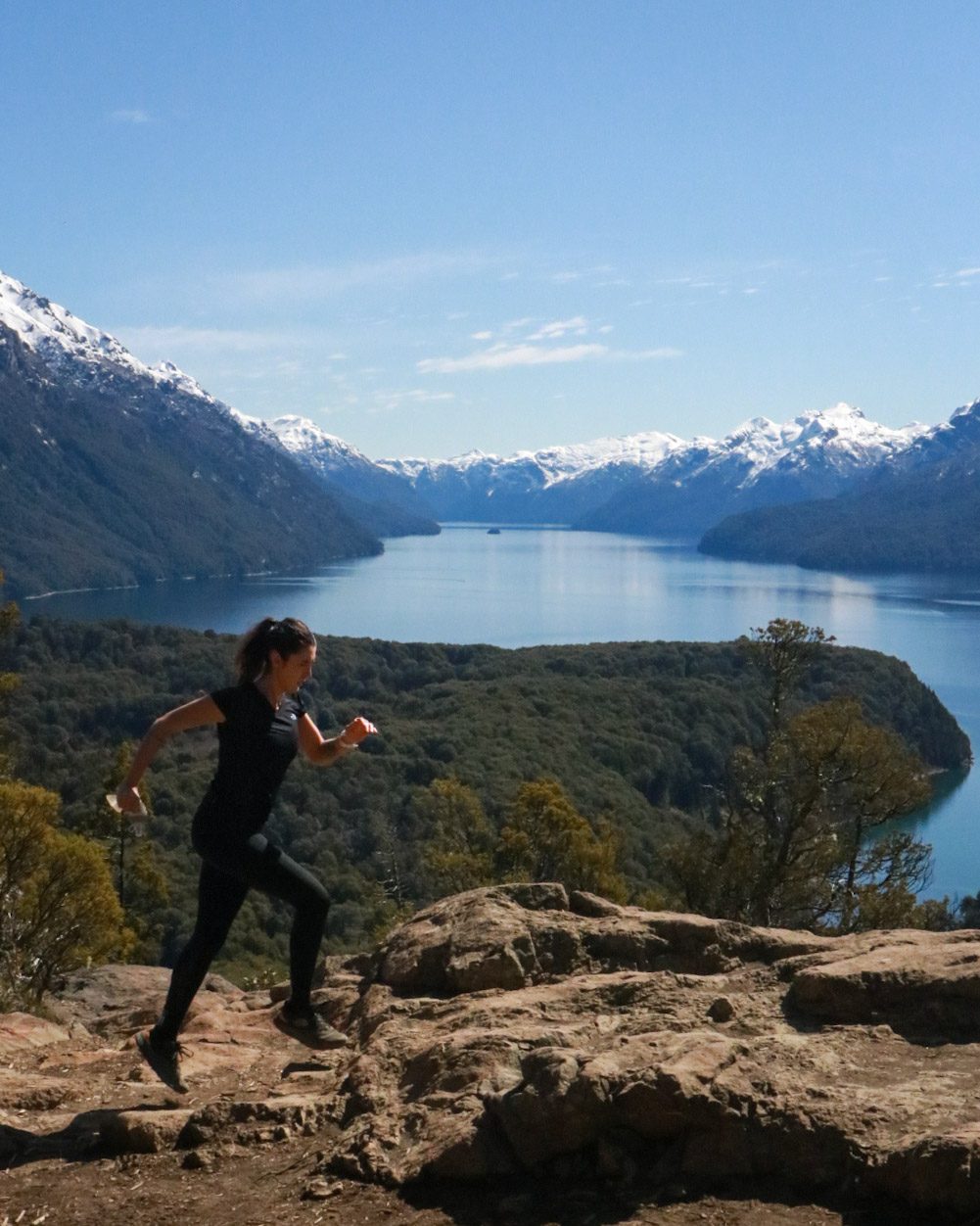 mulher correndo em trilha com lago e montanhas nevadas ao fundo em Bariloche