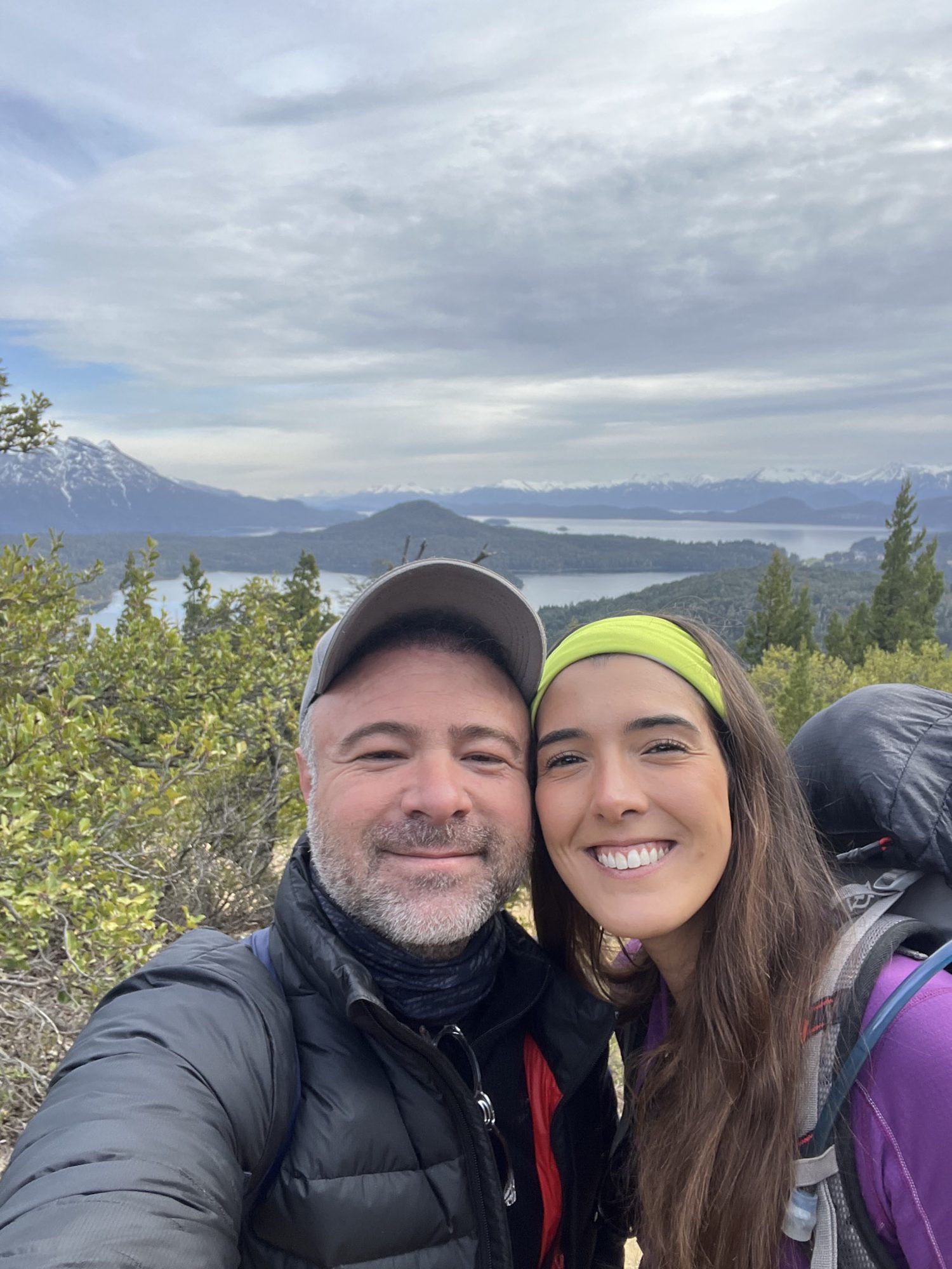 casal sorri em selfie no alto de trilha com lago e montanhas ao fundo em Bariloche