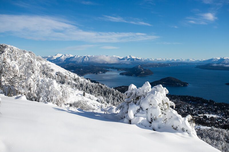 Mulher em selfie no topo de montanha rochosa com neve e lago ao fundo em Bariloche