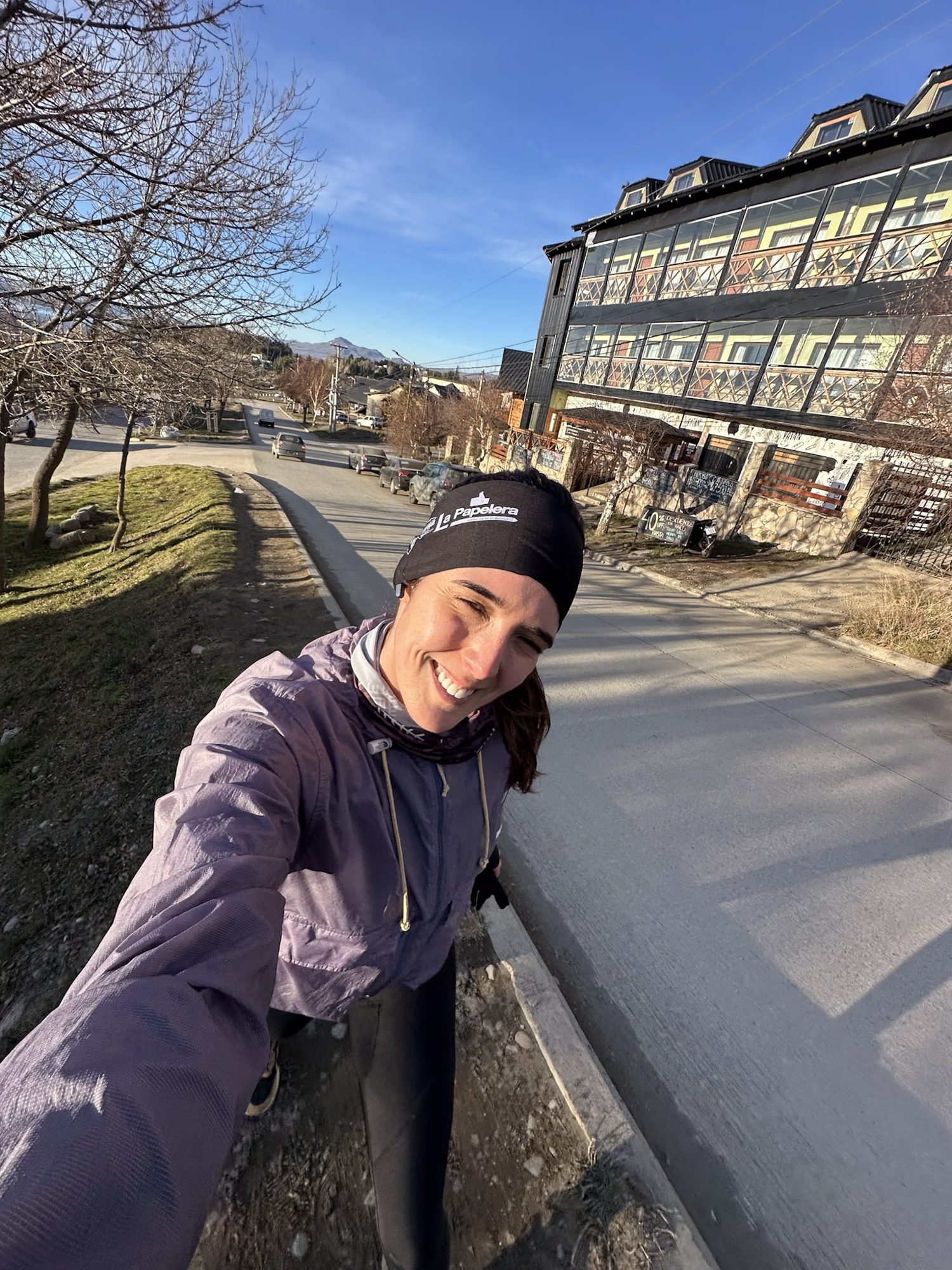Corredora sorridente tirando selfie em dia ensolarado nas ruas de Bariloche, com casas e montanhas ao fundo.