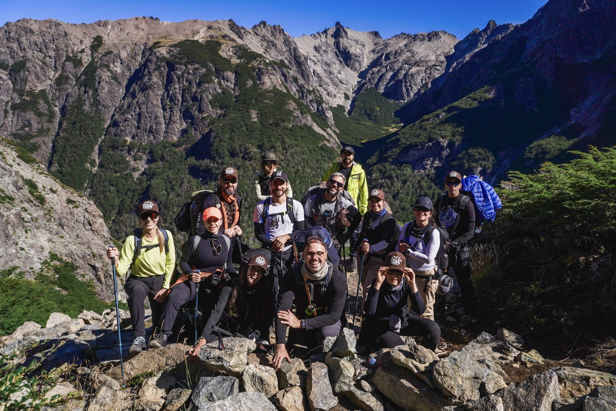 Grupo de trekkers posando em mirante rochoso durante trilha na Patagônia, com montanhas e vales verdes ao fundo.