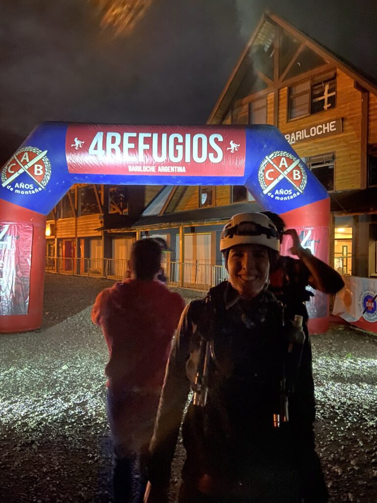 Corredora sorrindo sob a chuva na chegada da 4 Refugios Bariloche, à noite, com o arco inflável iluminado ao fundo.