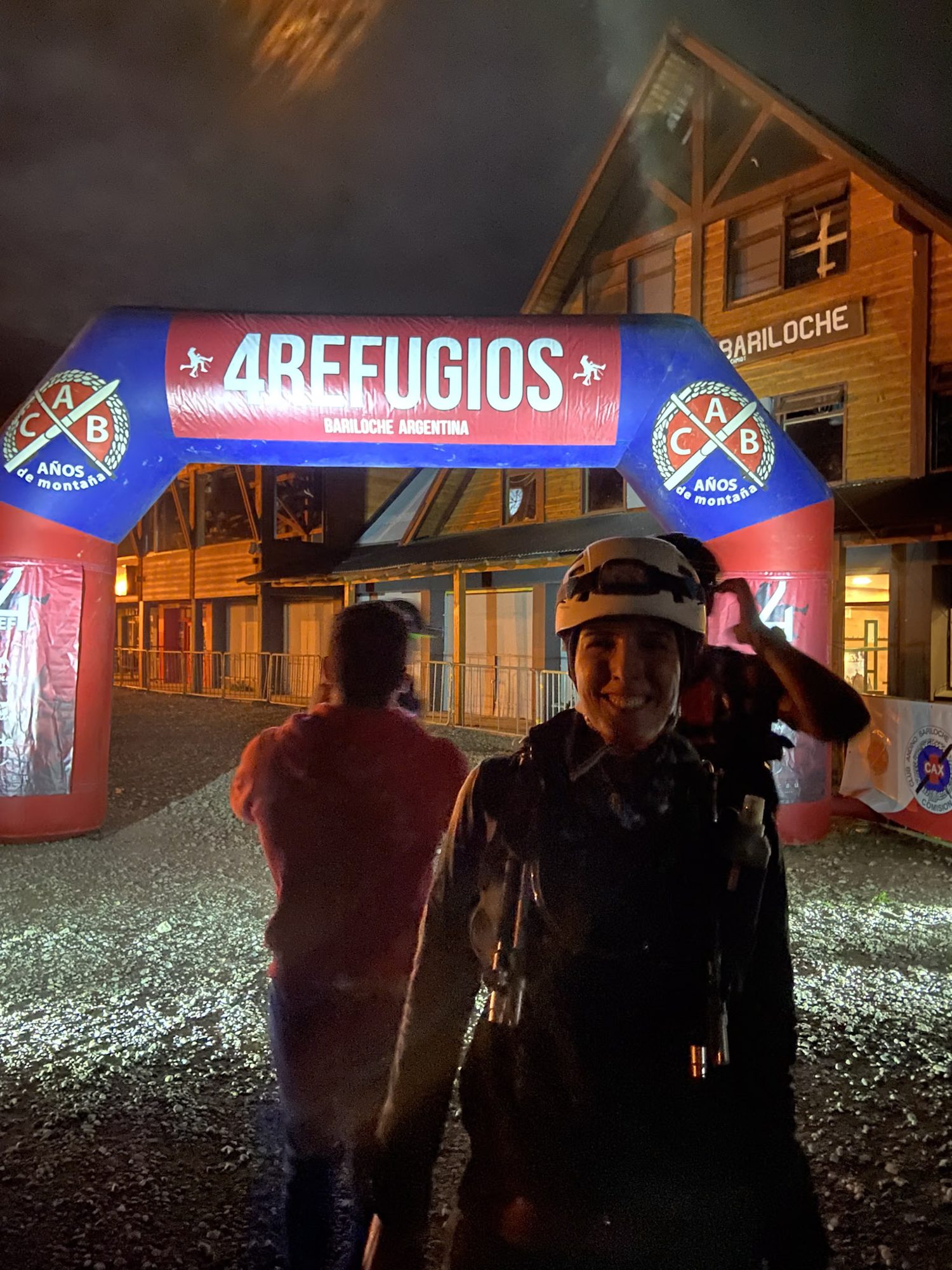 Corredora sorrindo sob a chuva na chegada da 4 Refugios Bariloche, à noite, com o arco inflável iluminado ao fundo.