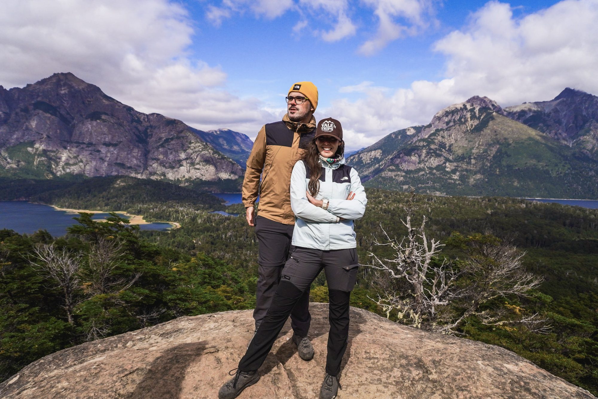 Casal no topo de um mirante na Patagônia argentina, com lago e montanhas ao fundo durante trekking em Bariloche.
