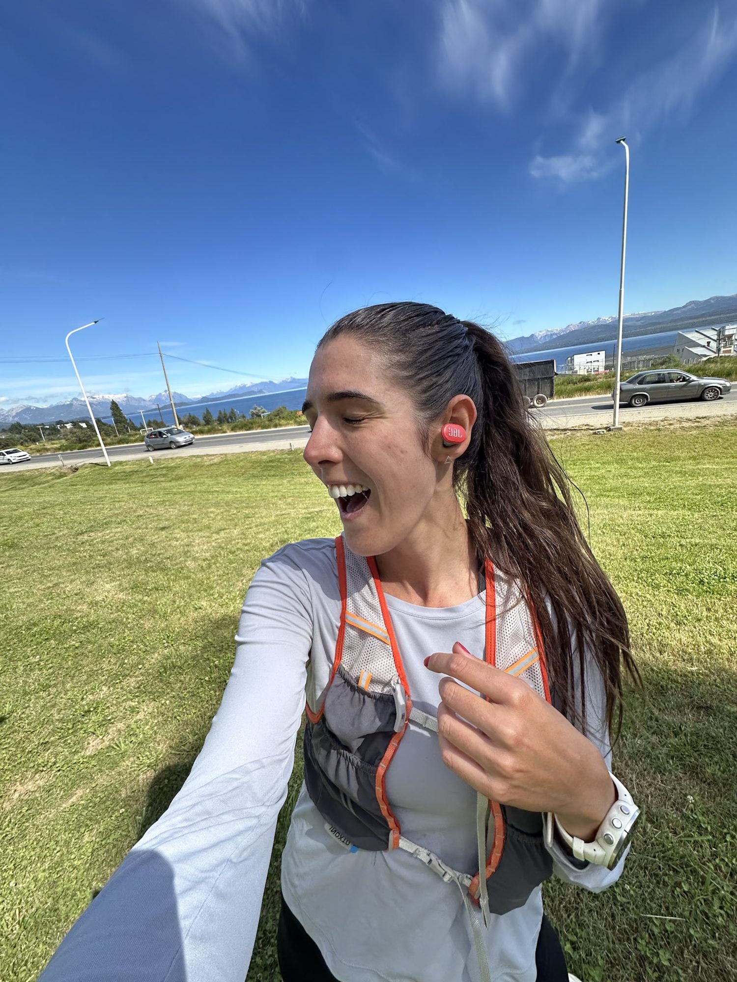 Corredora sorrindo com colete de hidratação após treino em Bariloche, com os Andes ao fundo sob céu azul.