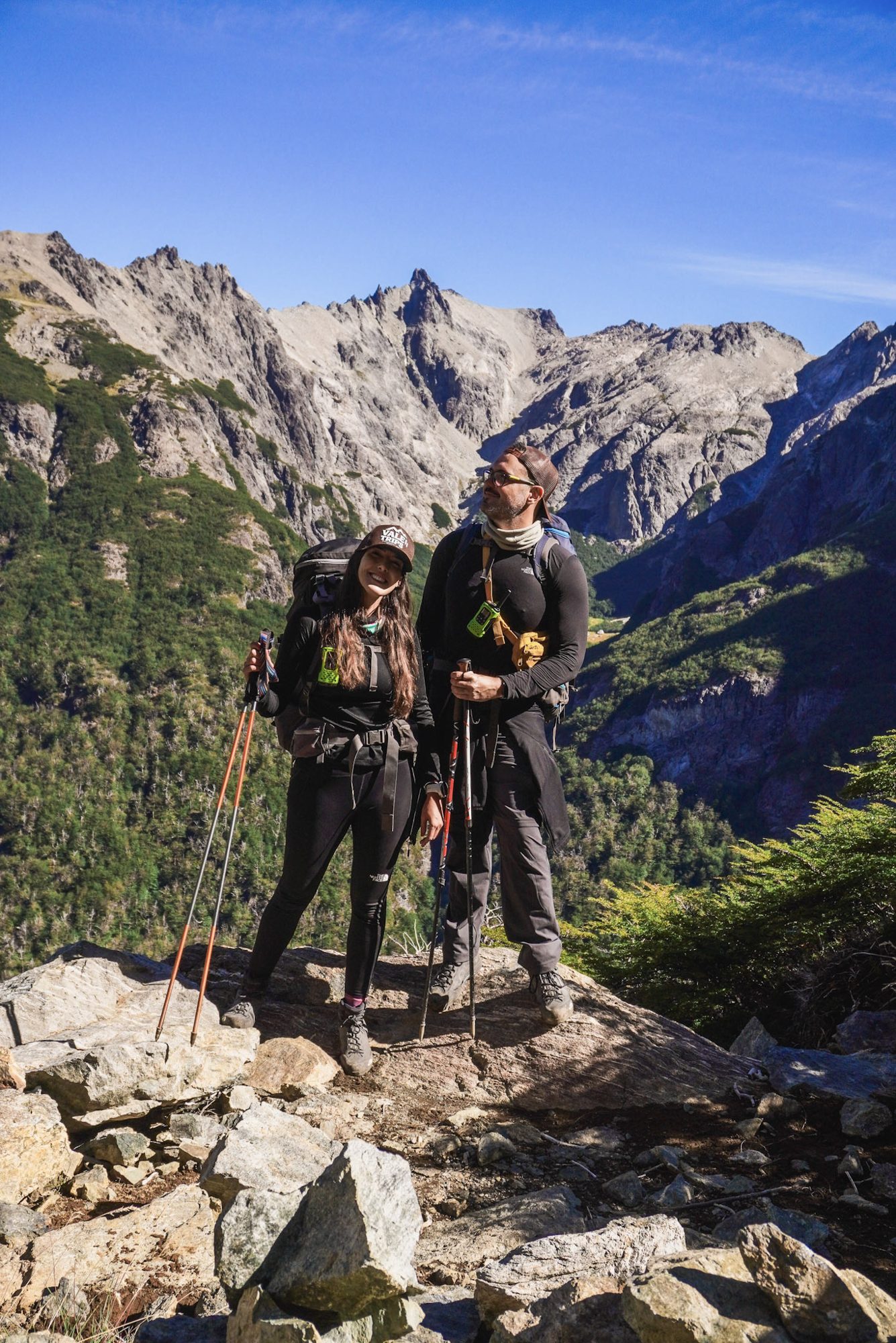Casal com mochilas e bastões de trekking posando em mirante rochoso com picos da Patagônia ao fundo.