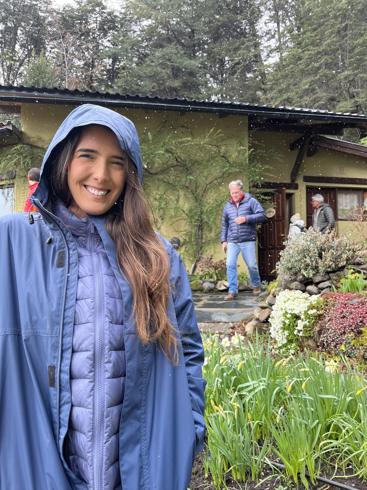 Bariloche na Primavera: Jovem sorrindo com capa de chuva azul em frente a casa com jardim florido em Bariloche - Vale Liberdade
