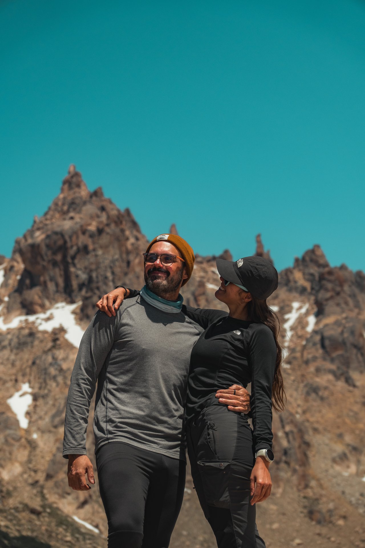 Casal em trekking com picos nevados ao fundo, reforçando como altitude e vento deixam a trilha mais fria.