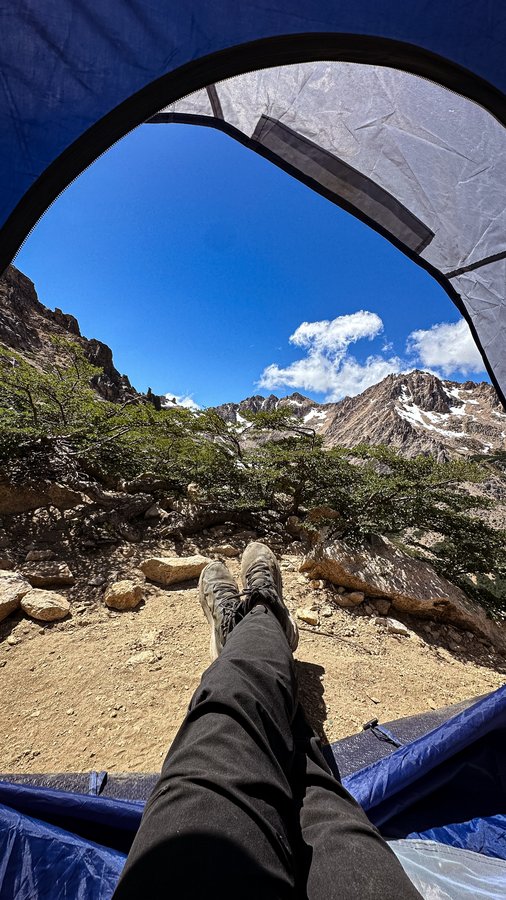Vista interna de barraca com pés de trekker em primeiro plano e montanhas com picos rochosos ao fundo