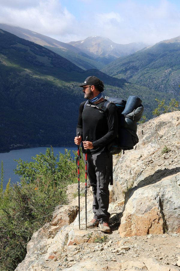 Homem em trek com mochila e bastões observando o vale com lago ao fundo
