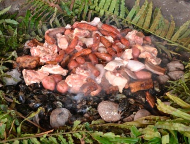Ingredientes do curanto em Bariloche sobre pedras quentes, com carnes, linguiças, batatas e folhas grandes ao redor.