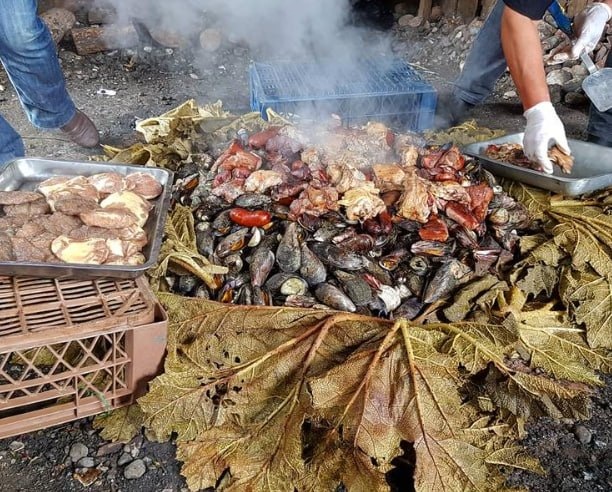 Destape do curanto em Colonia Suiza, com vapor saindo, carnes expostas e folhas grandes abertas ao redor do preparo.