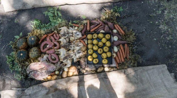 Curanto em Bariloche sendo montado no chão, com linguiças, legumes e folhas ao redor do preparo.