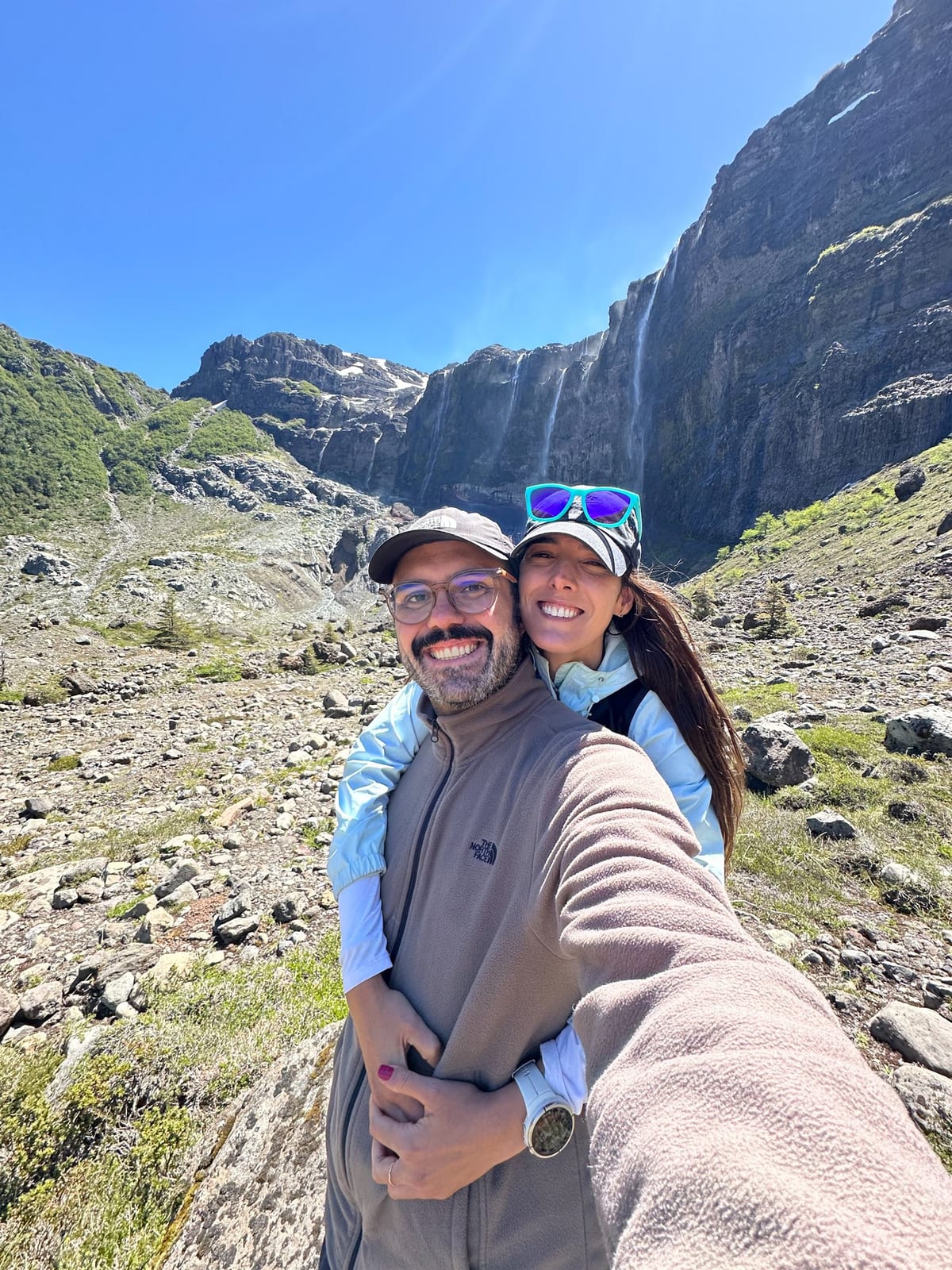 Gabriel e Dominique em trilha de verão em Bariloche, com cachoeira e paredão rochoso ao fundo.