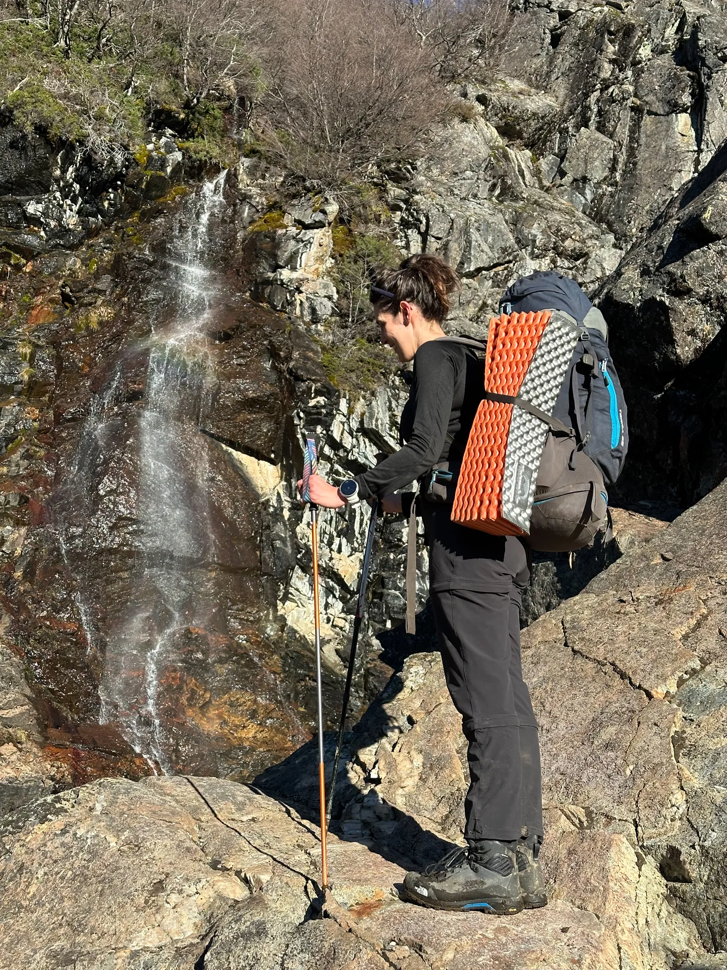 bariloche em abril: Pessoa fazendo trekking com mochila e bastão ao lado de uma cascata entre rochas em Bariloche, Patagônia argentina