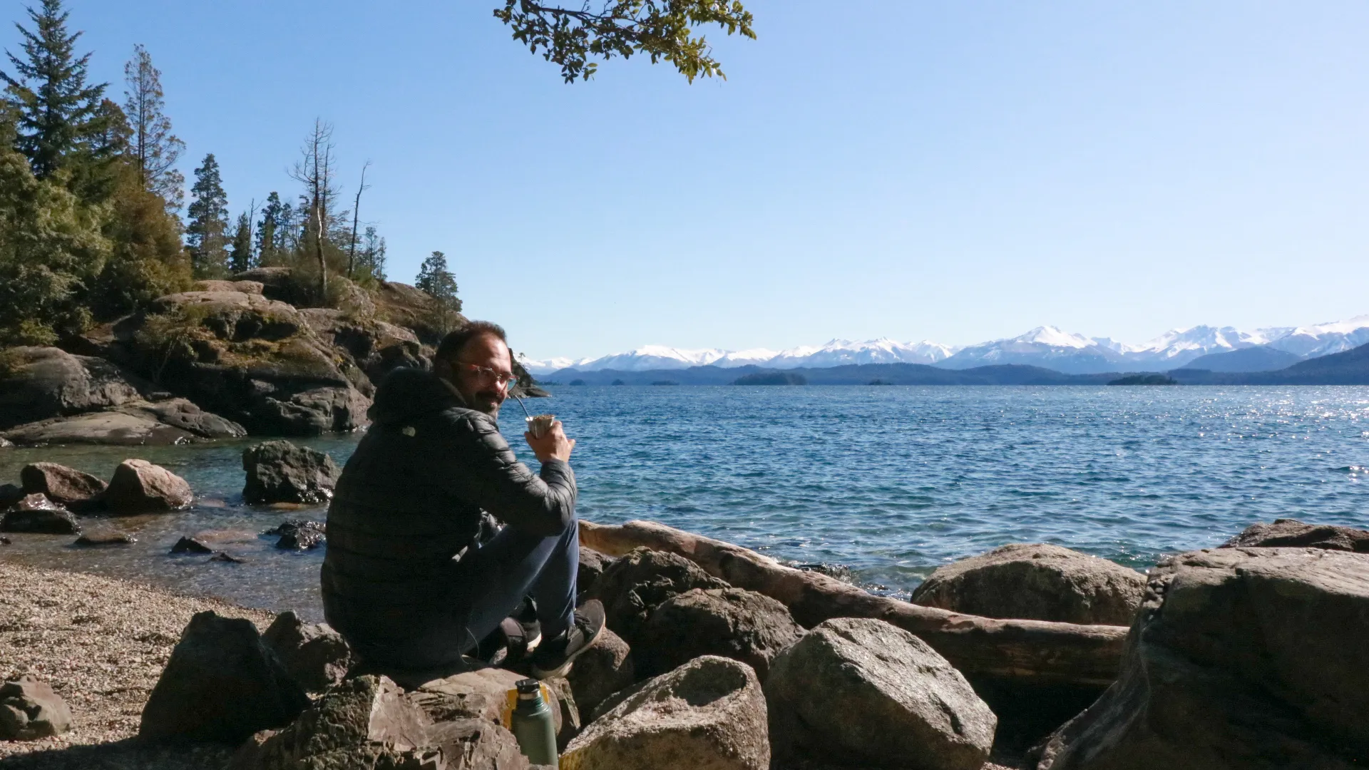 bariloche em abril: Viajante tomando mate nas pedras à beira do Lago Nahuel Huapi em Bariloche, com montanhas nevadas dos Andes ao fundo