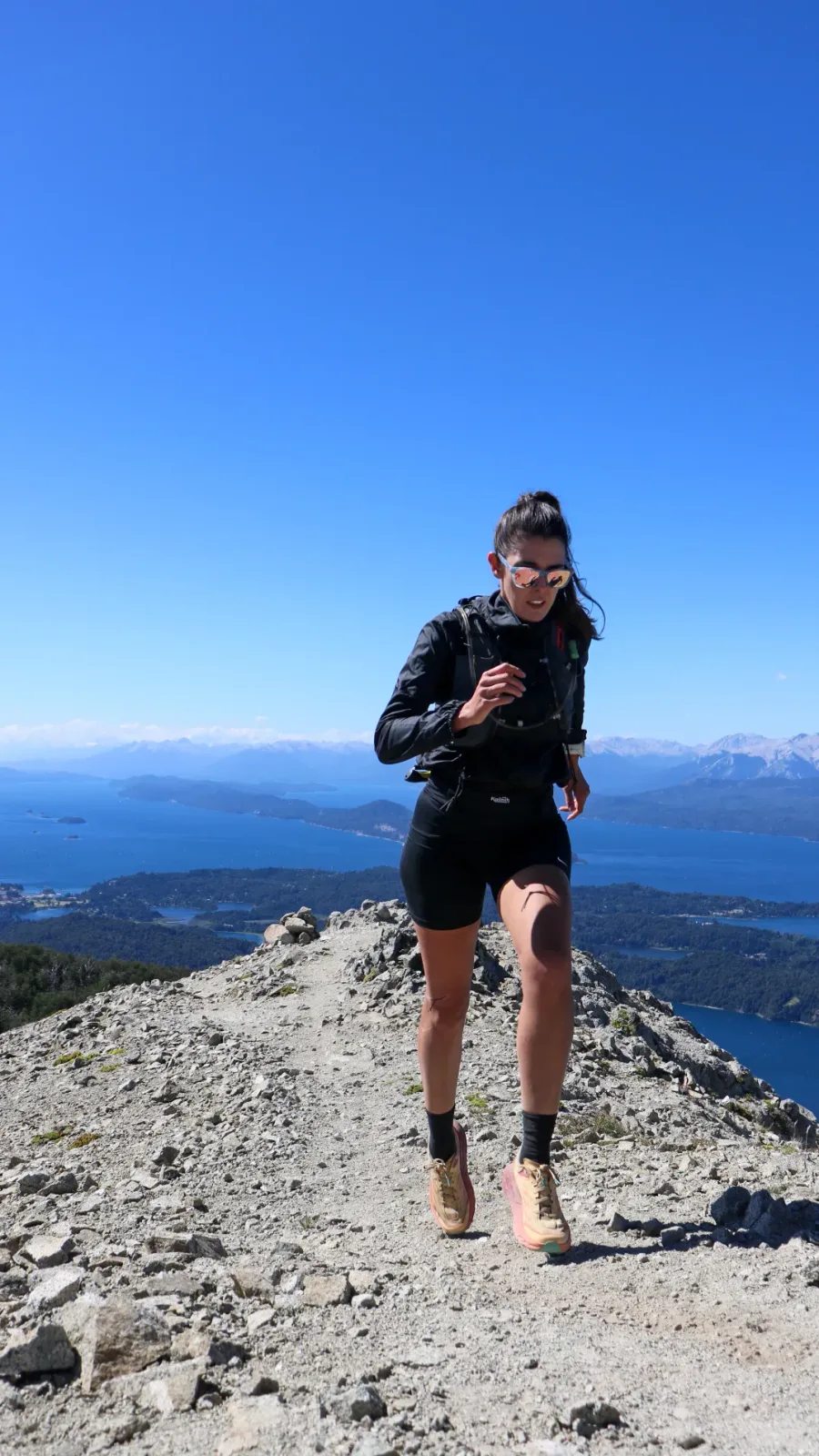 Bariloche em Fevereiro: Mulher praticando trail running no cume de uma montanha em Bariloche com vista panorâmica do Lago Nahuel Huapi e os Andes