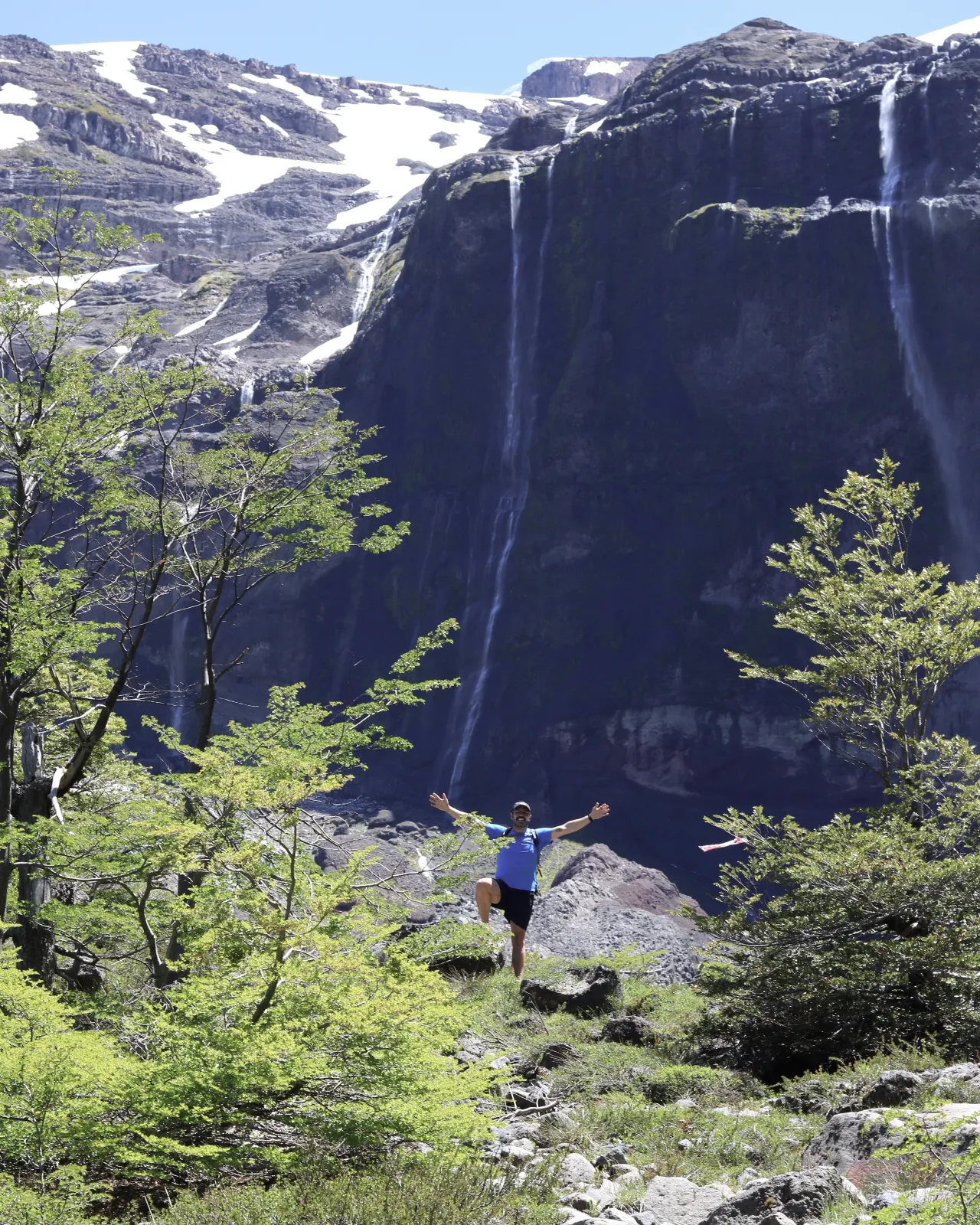 bariloche em janeiro: Pessoa com braços abertos em trilha diante de grande cascata e montanha com neve em Bariloche, Patagônia argentina