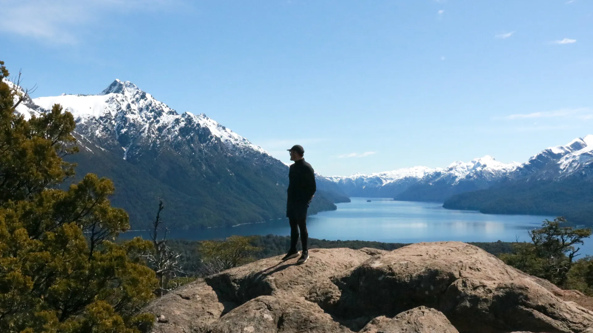 bariloche em maio: Viajante no alto de uma rocha contemplando o lago e os picos nevados da Patagônia argentina em Bariloche