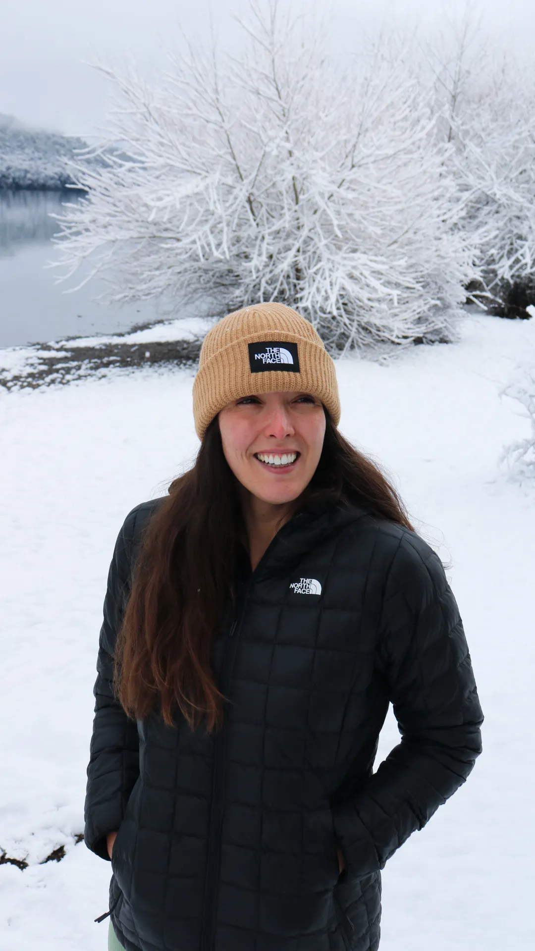 bariloche em maio: Mulher com jaqueta e gorro The North Face sorrindo na neve em Bariloche, com lago e árvores cobertas de geada ao fundo