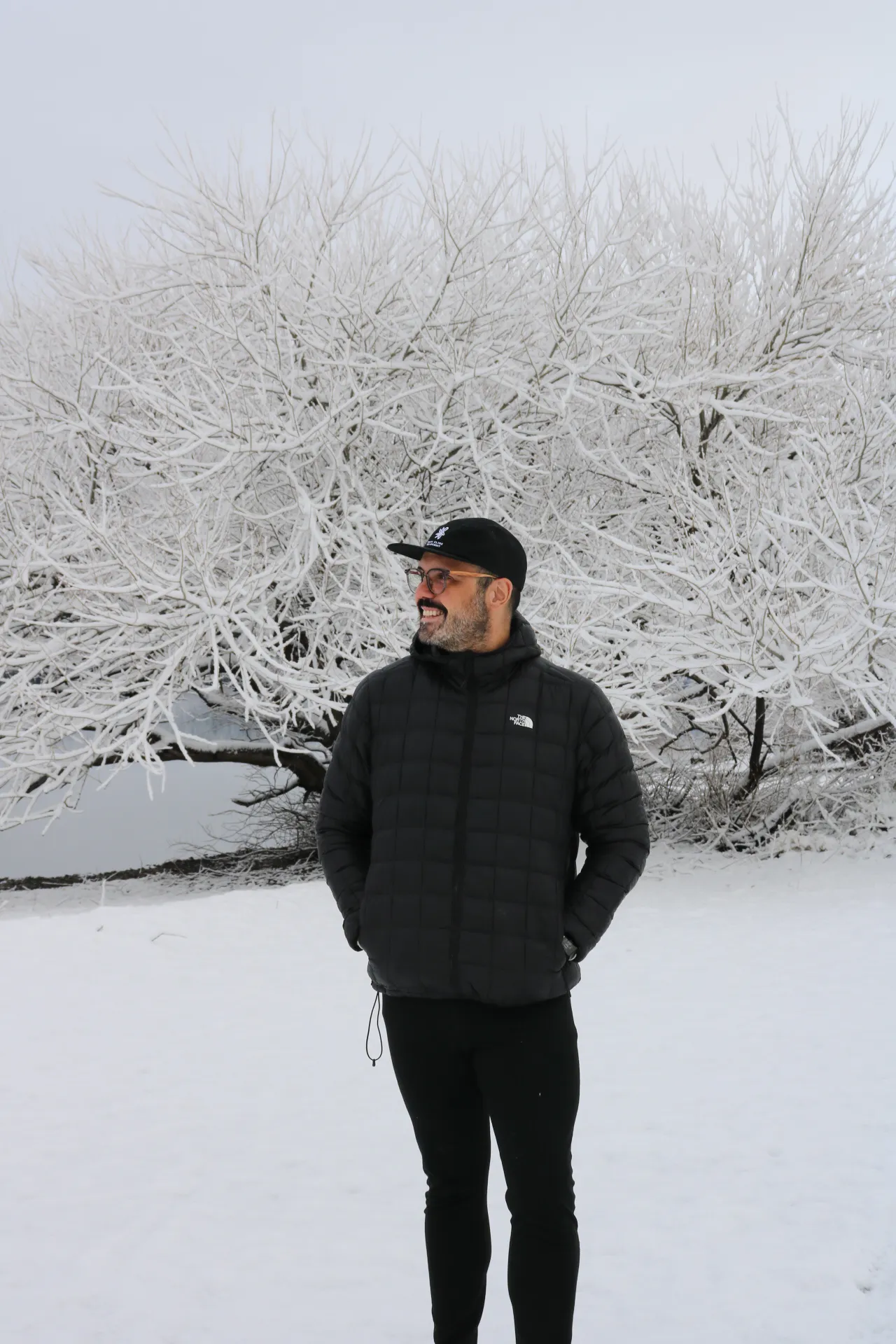 bariloche em maio: Homem com casaco preto The North Face posando na neve em Bariloche, Patagônia argentina, com árvores cobertas de geada ao fundo