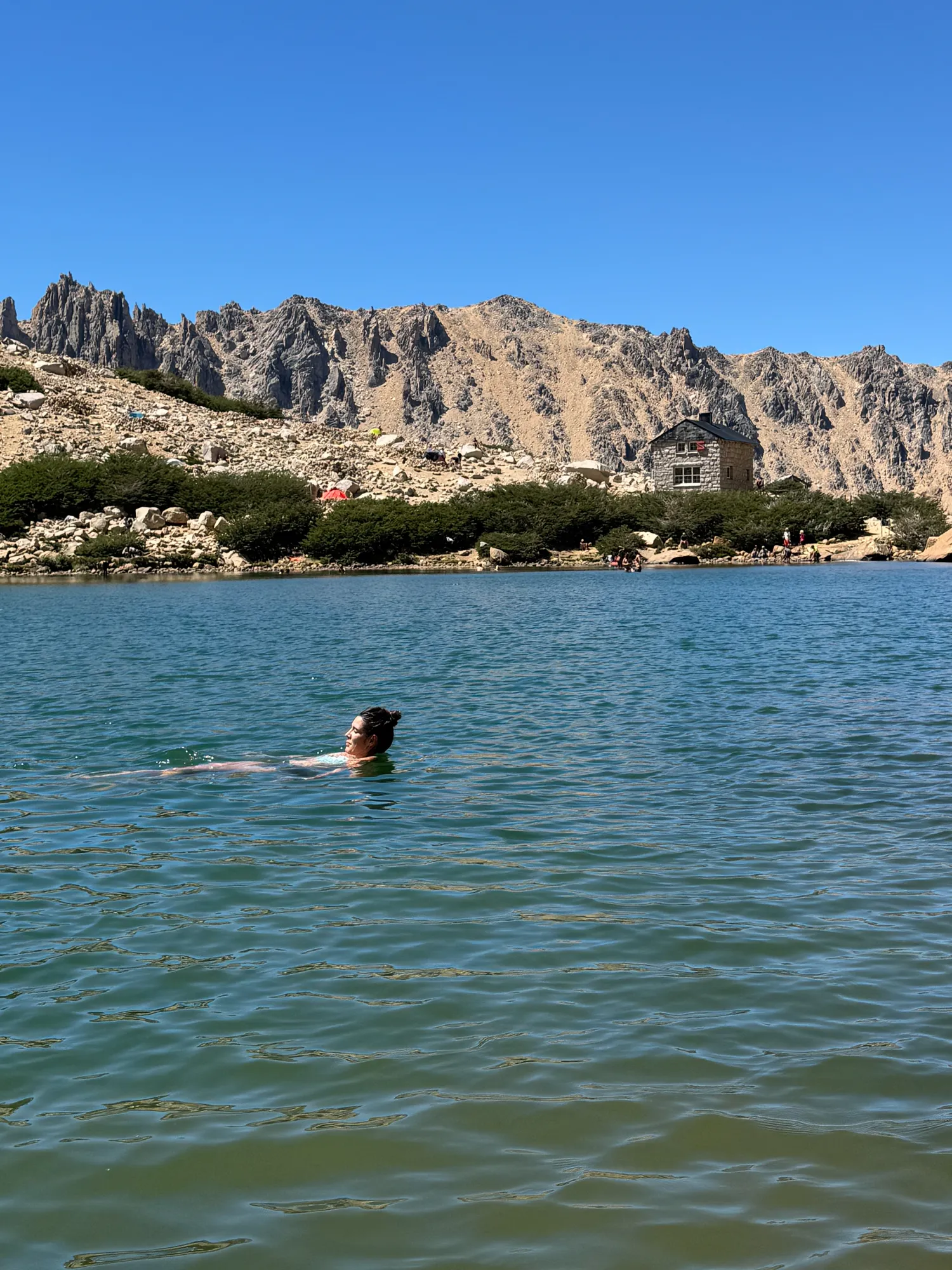 bariloche em março: Pessoa nadando no Lago Toncek com o Refugio Frey e as torres de granito do Cerro Catedral ao fundo, Bariloche