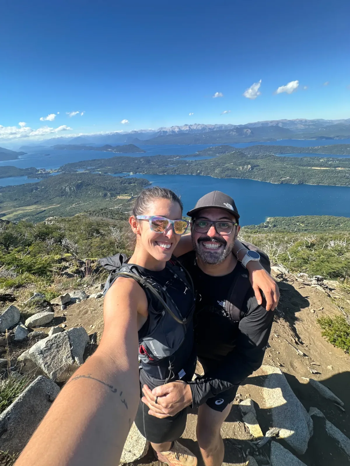 cerro campanario: Casal sorrindo no topo de trilha em Bariloche com vista do Lago Nahuel Huapi e montanhas da Patagônia ao fundo