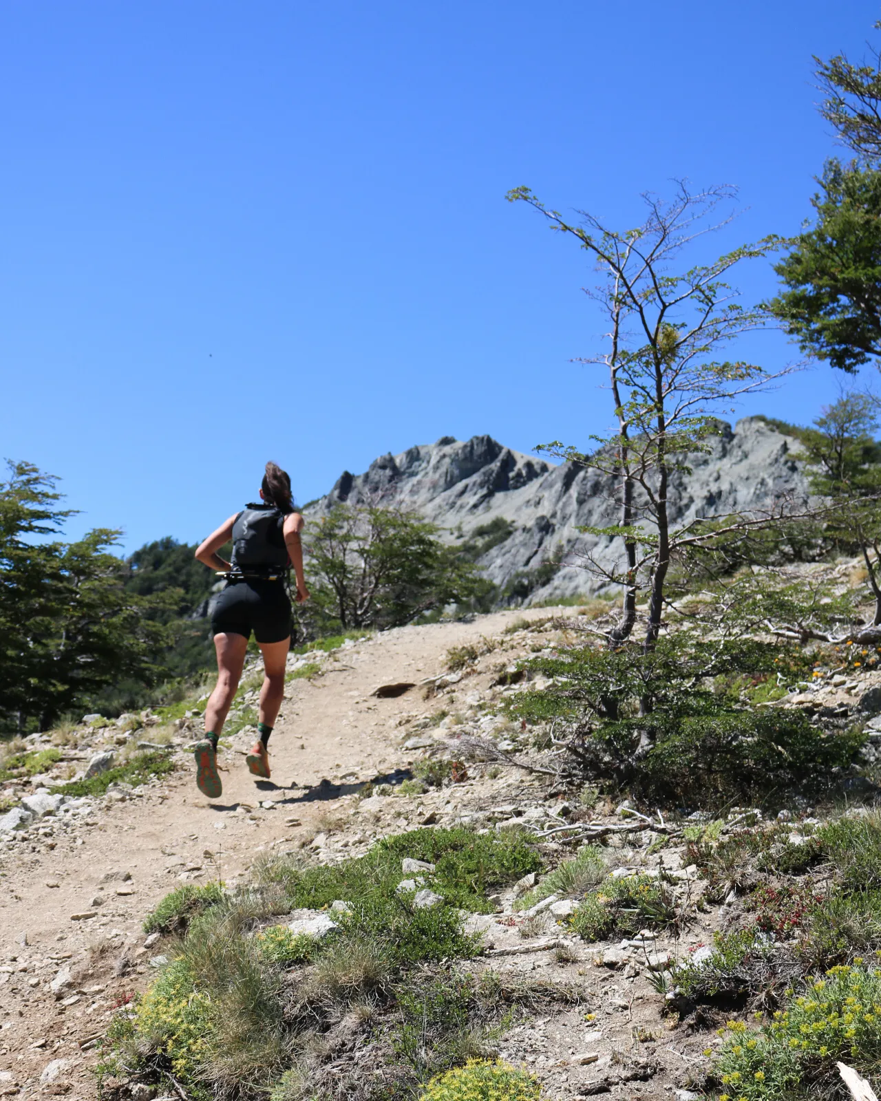 cerro campanario: Mulher praticando trail running em trilha de montanha em Bariloche, com picos rochosos e árvores nativas da Patagônia ao fundo