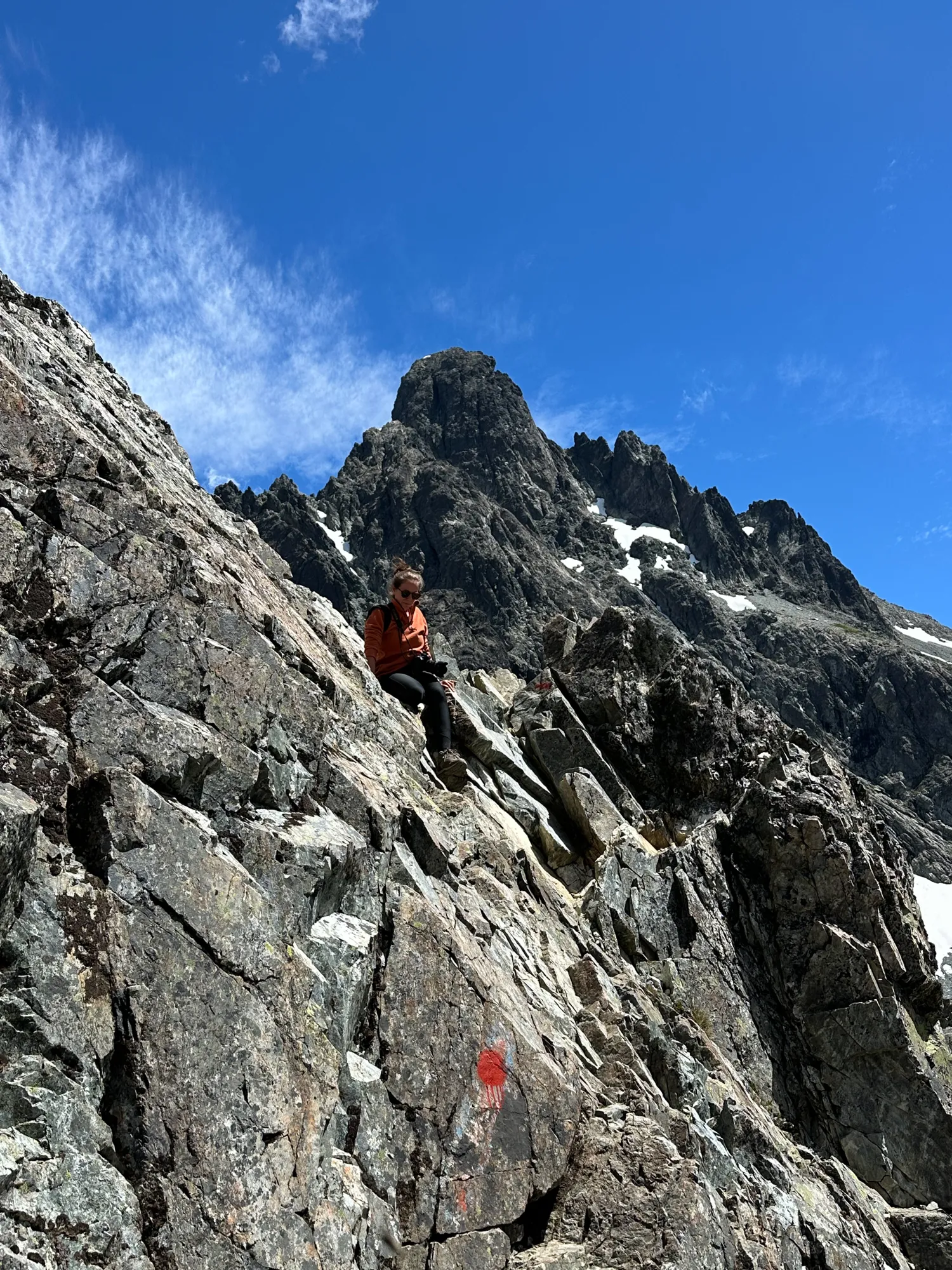 k42: Trilheiro descansando em trecho rochoso e exposto de trekking na Patagônia argentina, com picos graníticos e neve ao fundo em Bariloche
