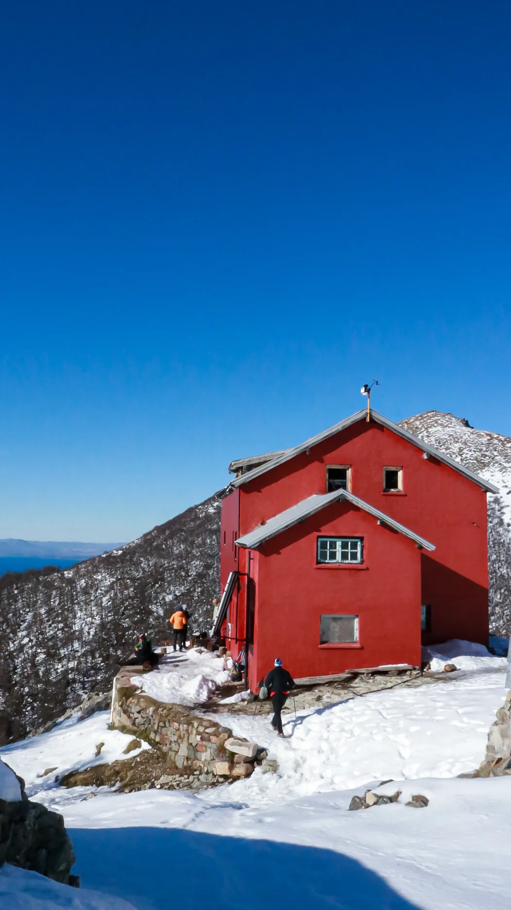 o que é trail run: Refúgio vermelho no topo nevado da serra de Bariloche com trilheiros e vista do lago Nahuel Huapi ao fundo