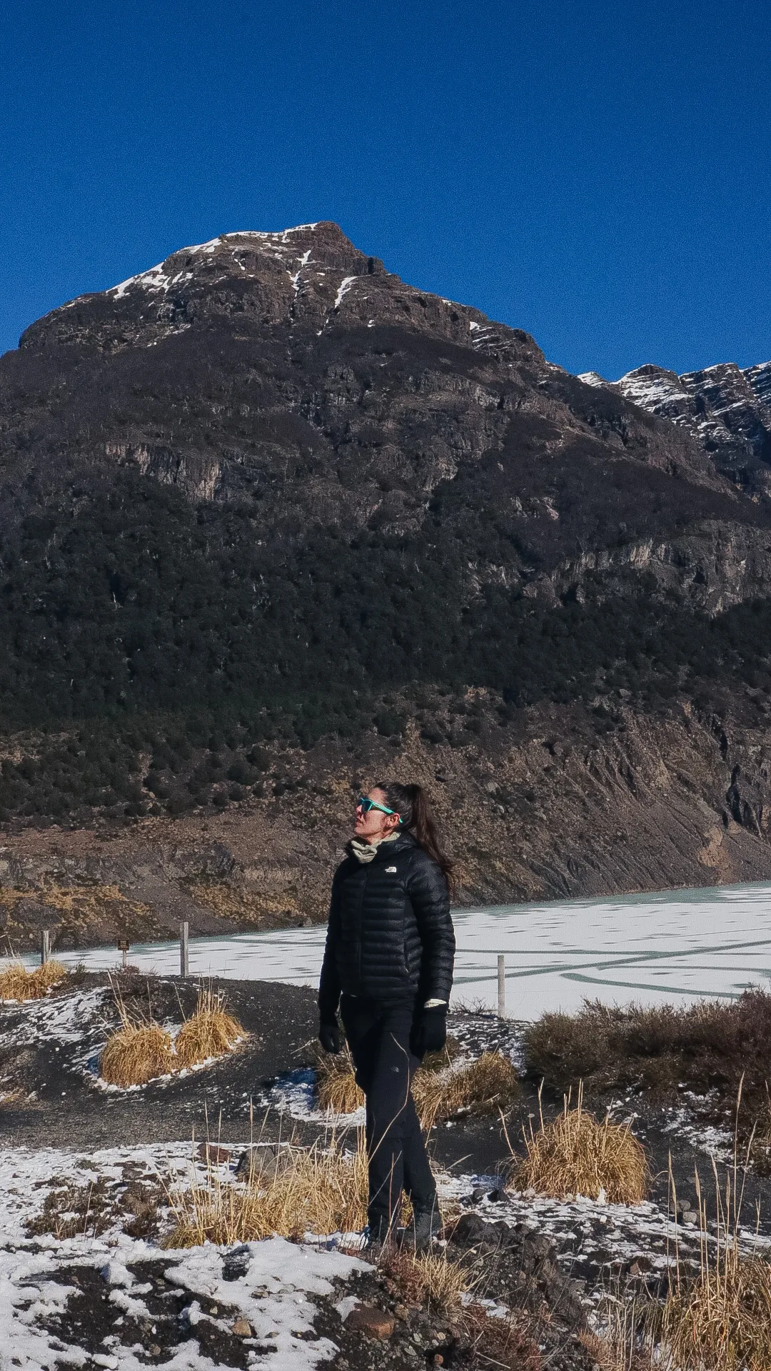 o que é trekking: Mulher com jaqueta preta The North Face em frente a lago congelado e montanha com neve na Patagônia argentina, Bariloche