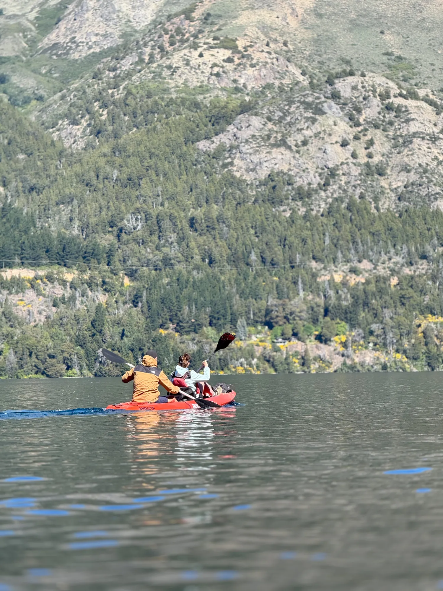 patagonia run: Casal remando caiaque vermelho no Lago Gutiérrez, com encosta arborizada ao fundo em Bariloche.