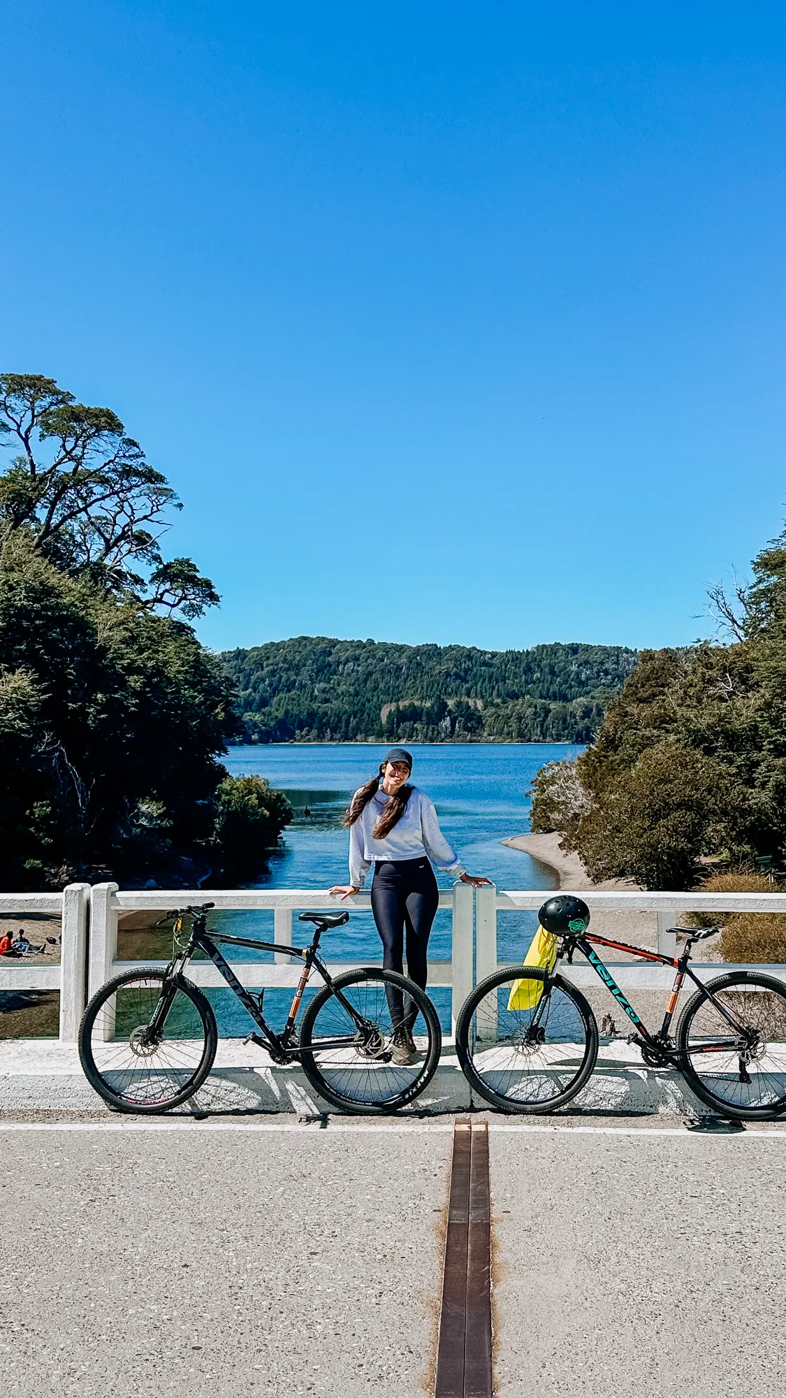 patagonia run: Mulher posando com duas bicicletas em ponte sobre lago em Bariloche, Patagônia argentina, com vegetação e céu azul ao fundo