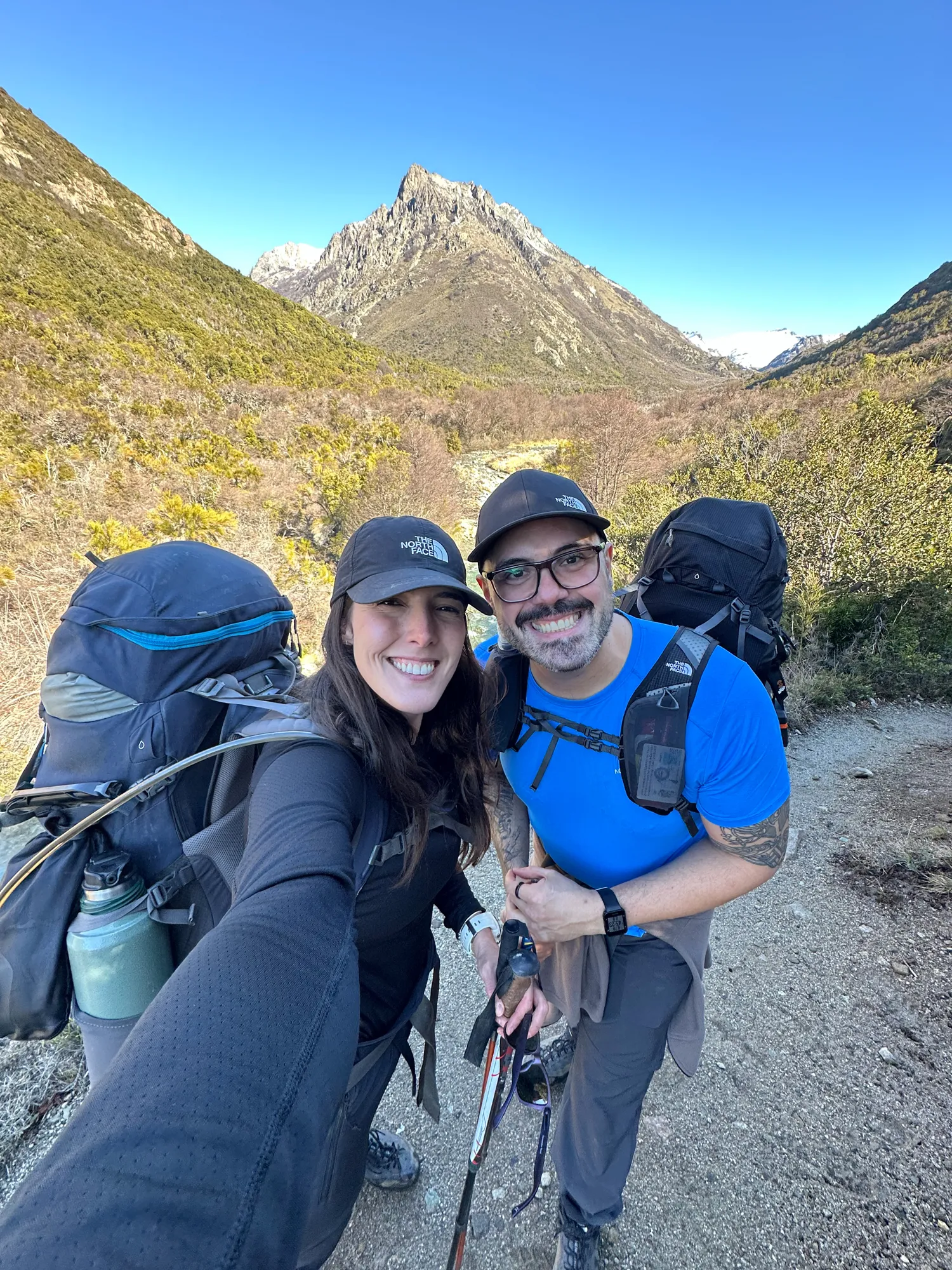 refugio jakob bariloche: Casal com mochilas de trilha sorri em selfie na trilha do Refúgio Jakob, com montanhas rochosas ao fundo em Bariloche.