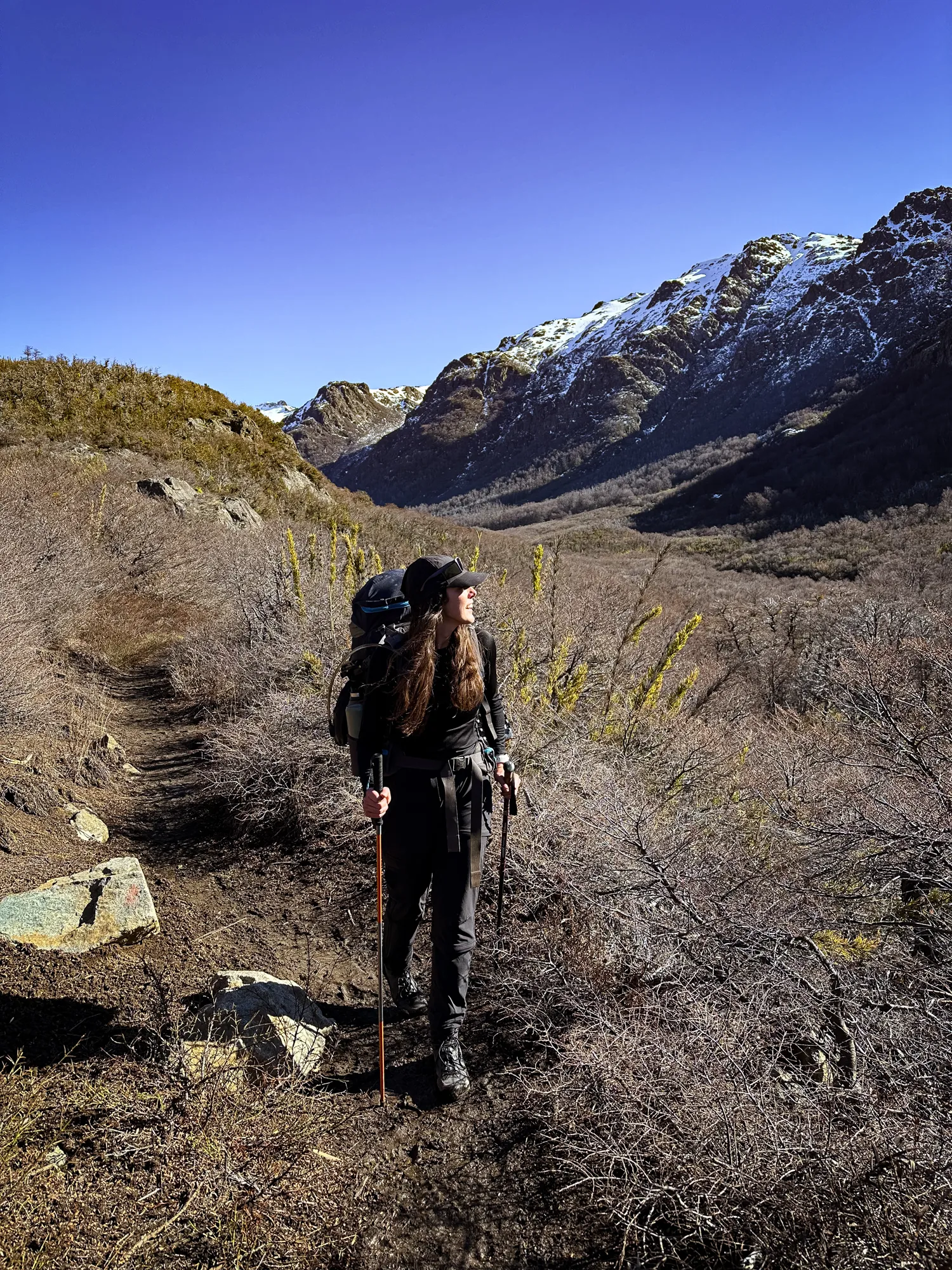 refugio jakob bariloche: Mulher com mochila e bastões caminha por trilha na trilha do Refúgio Jakob, com montanhas nevadas ao fundo em Bariloche.