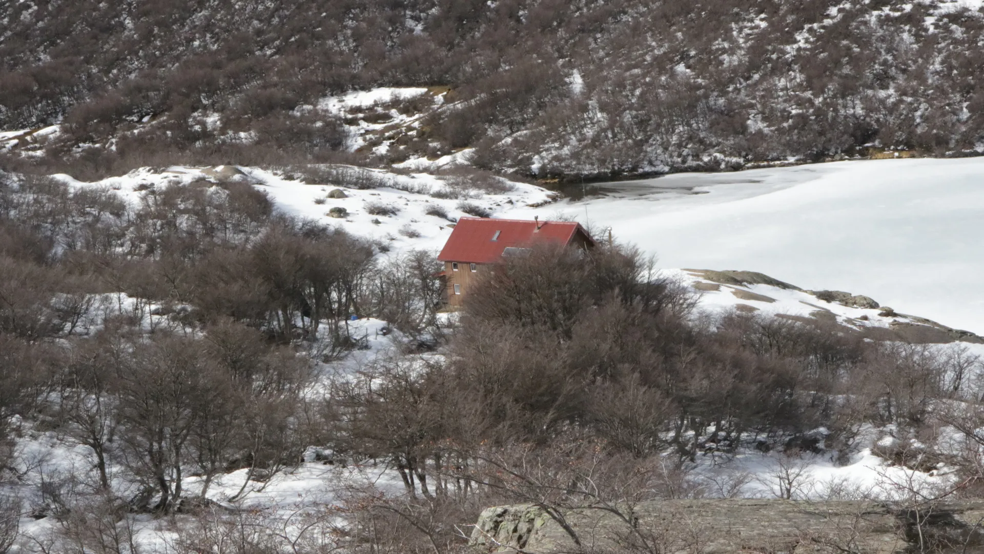 refugio jakob bariloche: Refúgio Jakob com telhado vermelho cercado por vegetação seca e neve, ao lado de uma lagoa congelada em Bariloche.
