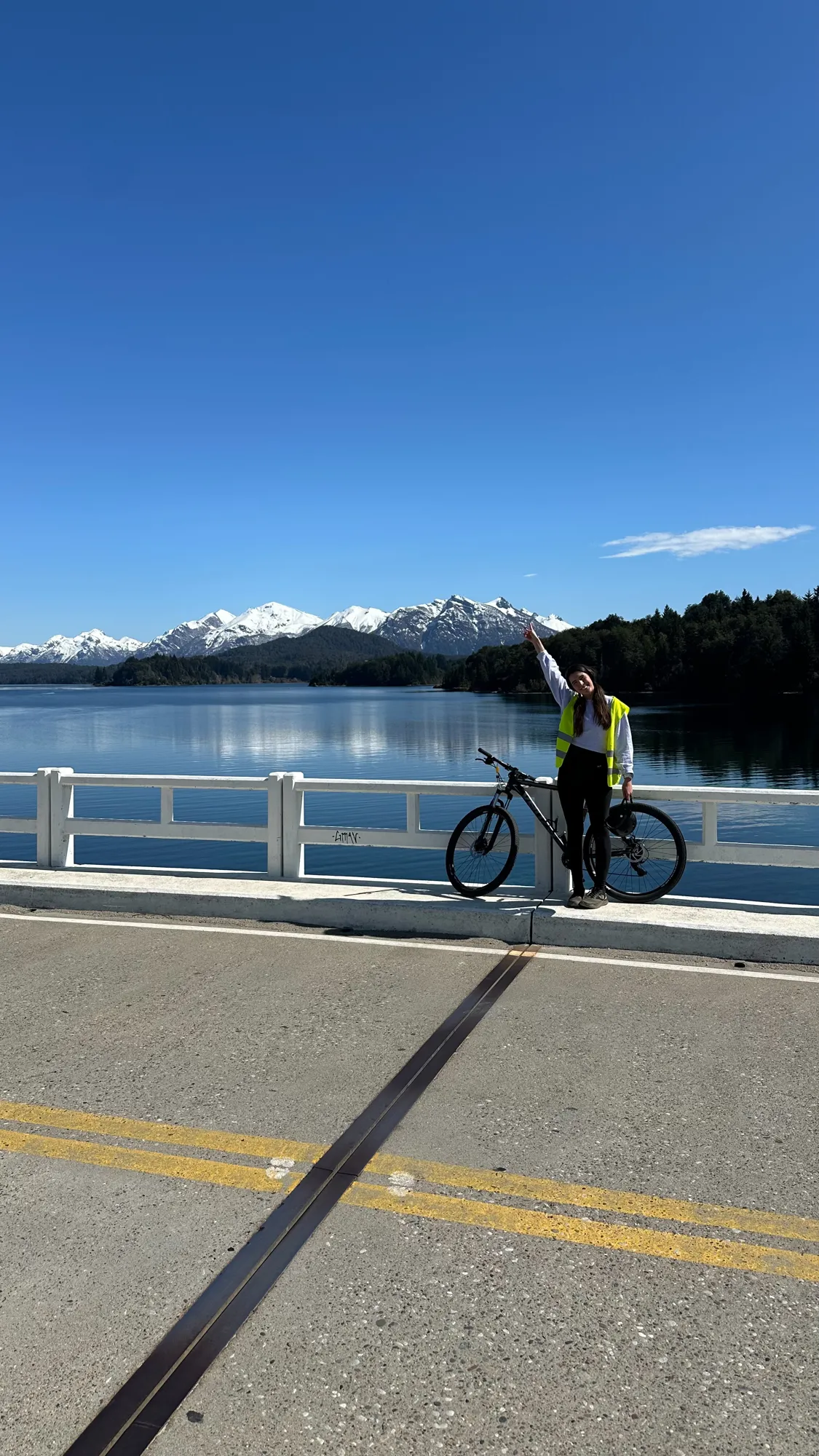 roteiro bariloche inverno: Ciclista posando com bicicleta em ponte sobre o Lago Nahuel Huapi com picos nevados ao fundo em Bariloche, Patagônia argentina