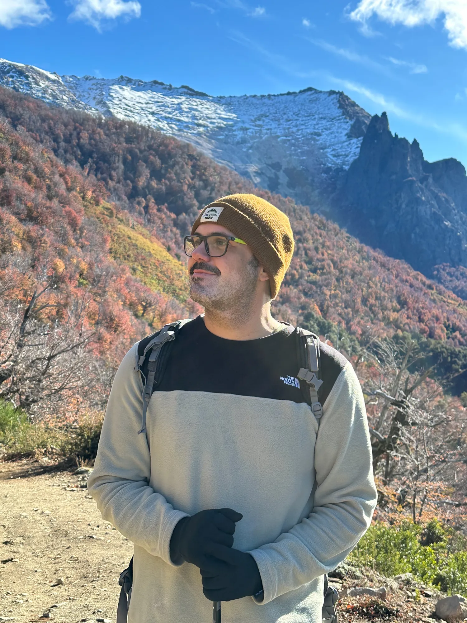 roteiro bariloche inverno: Trilheiro com mochila e touca em trilha de Bariloche com montanhas nevadas e lengas coloridas ao fundo no outono patagônico