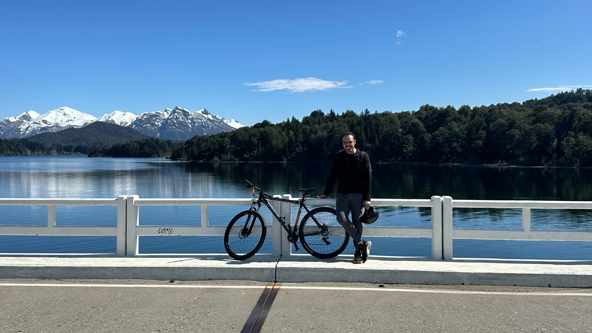 roteiro bariloche inverno: Homem com mountain bike em ponte sobre o Lago Nahuel Huapi em Bariloche, com montanhas nevadas dos Andes ao fundo