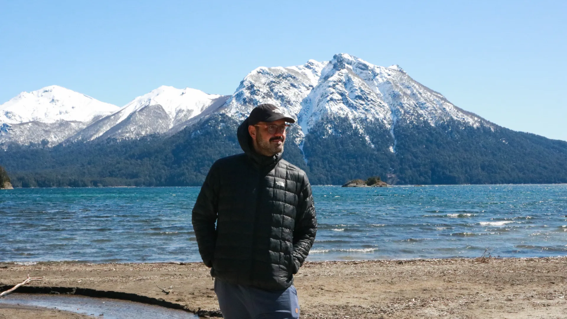 roteiro bariloche inverno: Homem na beira do lago Nahuel Huapi em Bariloche com montanhas nevadas e água turquesa ao fundo